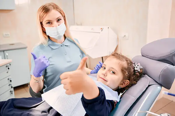 A happy little girl and a masked dental hygienist giving thumbs up together from the dental chair.